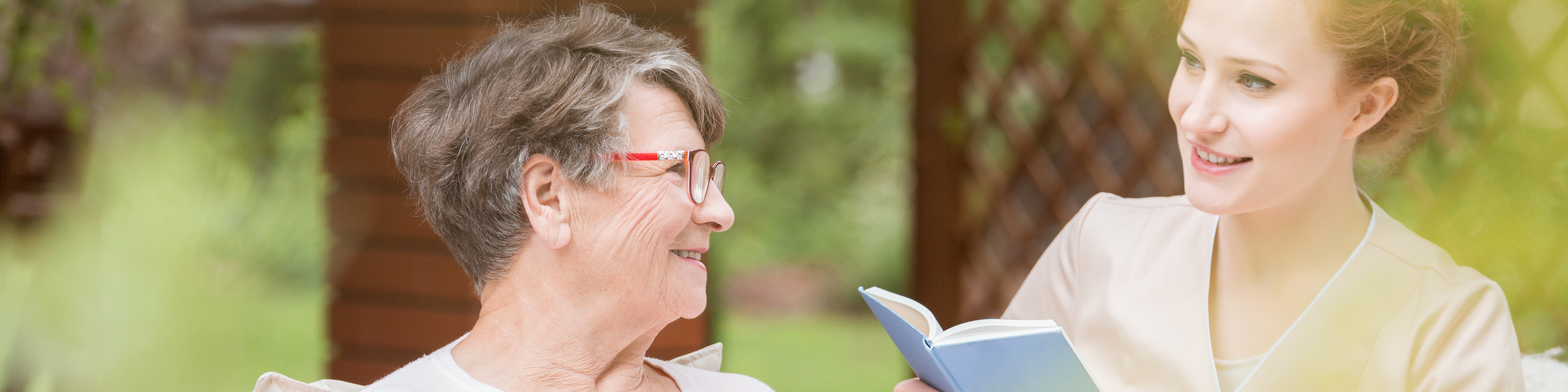 Eine Betreuerin liest einer älteren Dame, die eine Tasse in der Hand hält etwas aus einem Buch vor | © Photographee.eu - stock.adobe.com